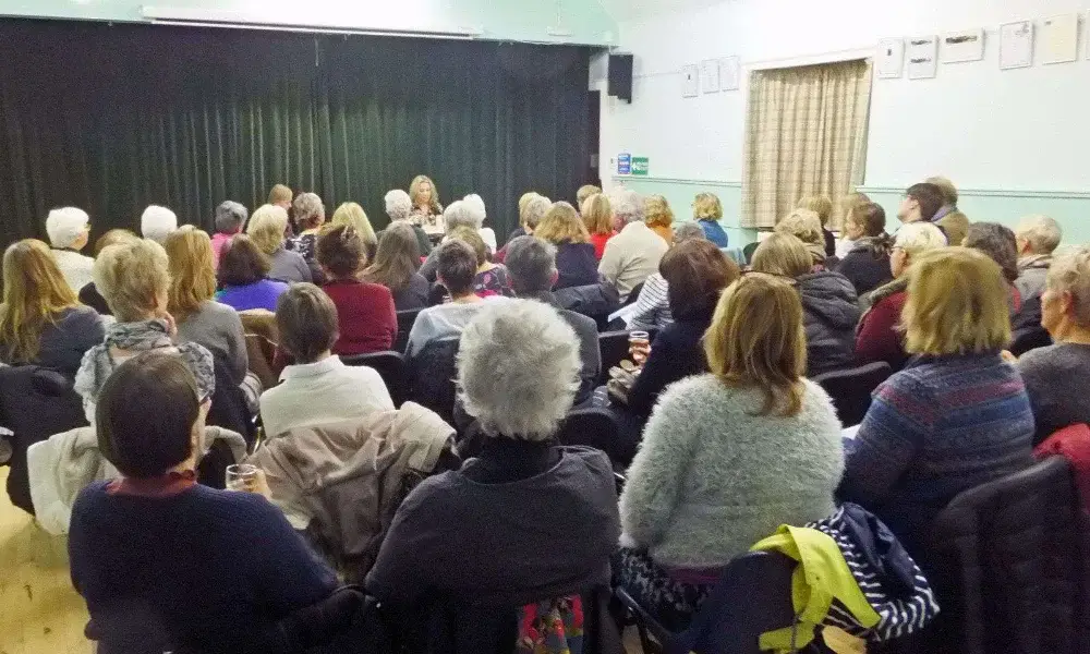 Audience seated in rows facing a stage with a green curtain in a light-walled room